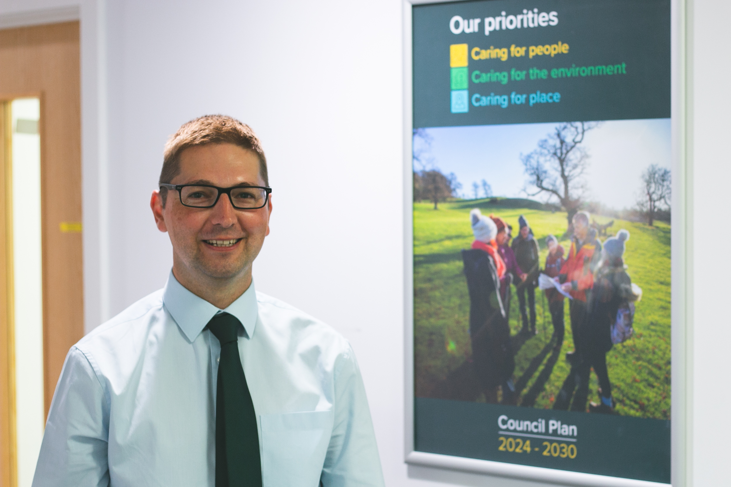 Cllr Richard Stanley smiling and standing in from of the Tewkesbury Borough Council Our Priorities image: text reads caring for people, environment and place.