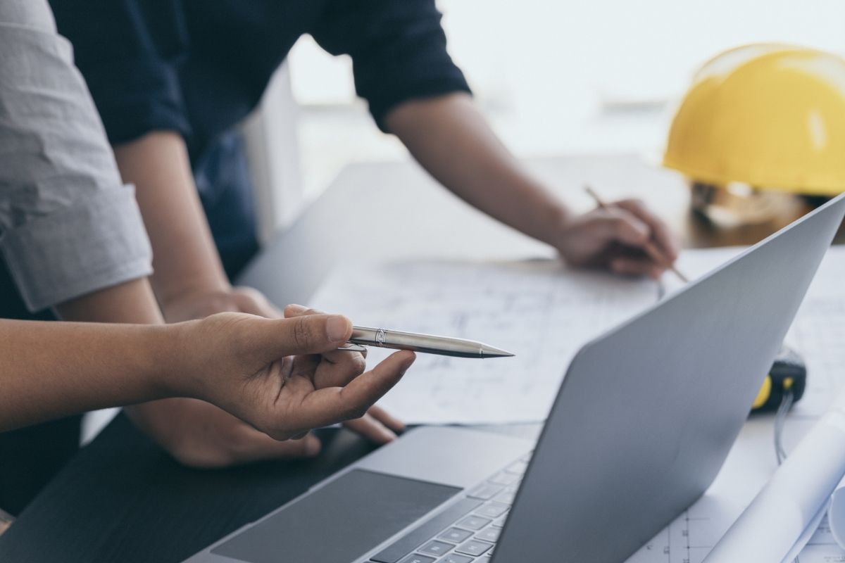Photo cropped closely to show two people leaning over planning documents and a laptop, with a bright yellow hard hat in the background