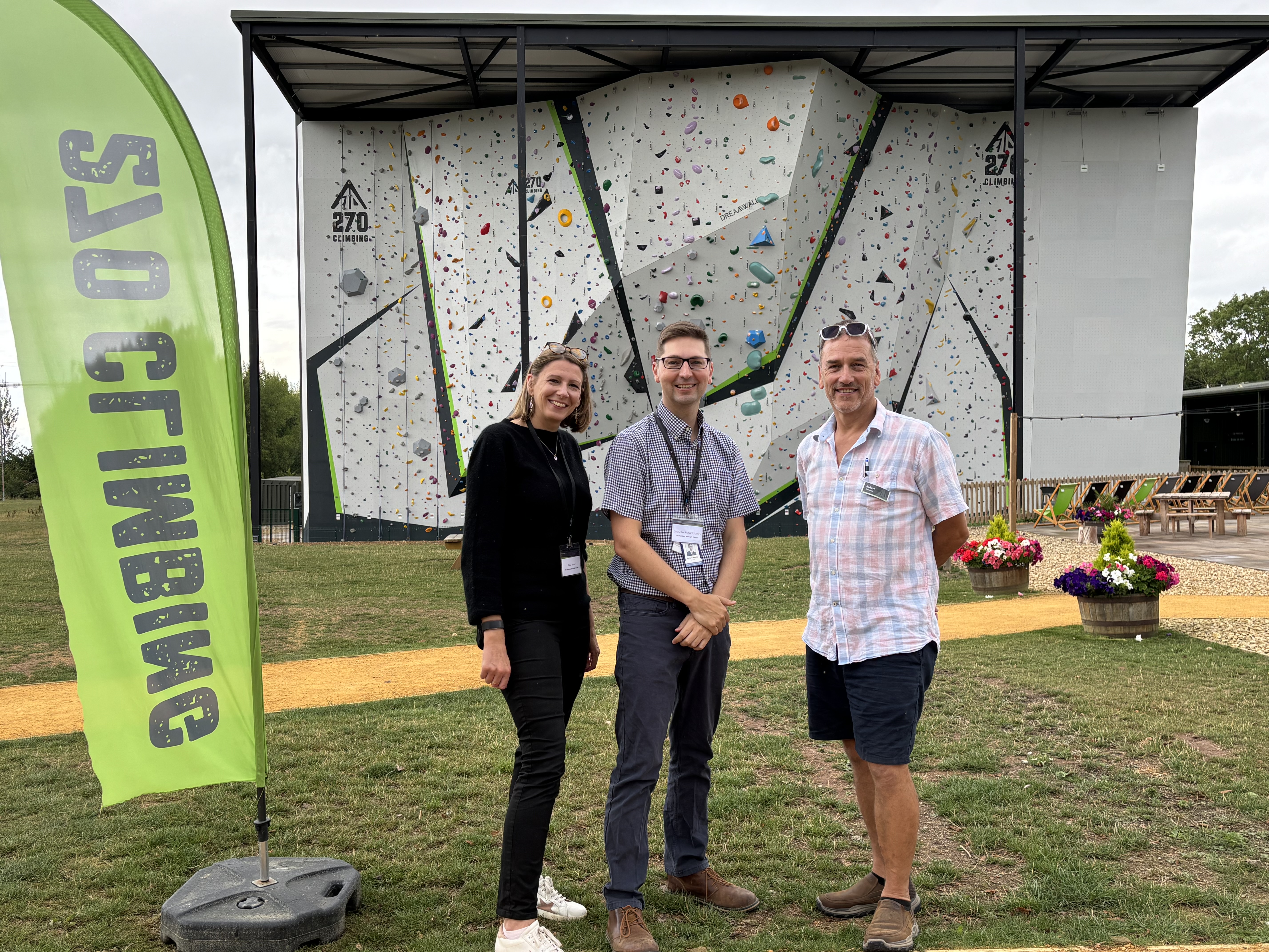 An image of 270 Climbing Park Olympic standard climbing wall in the background and in the foreground stands Katie Power Business growth and enterprise manager The Growth Hub - Tewkesbury, Leader Cllr Richard Stnaley and Lead member for Economic Development to the right is Robert Stevens 270 Climbing Park co-owner. flowered baskets are to the side along with a bright banner with 270 logo.