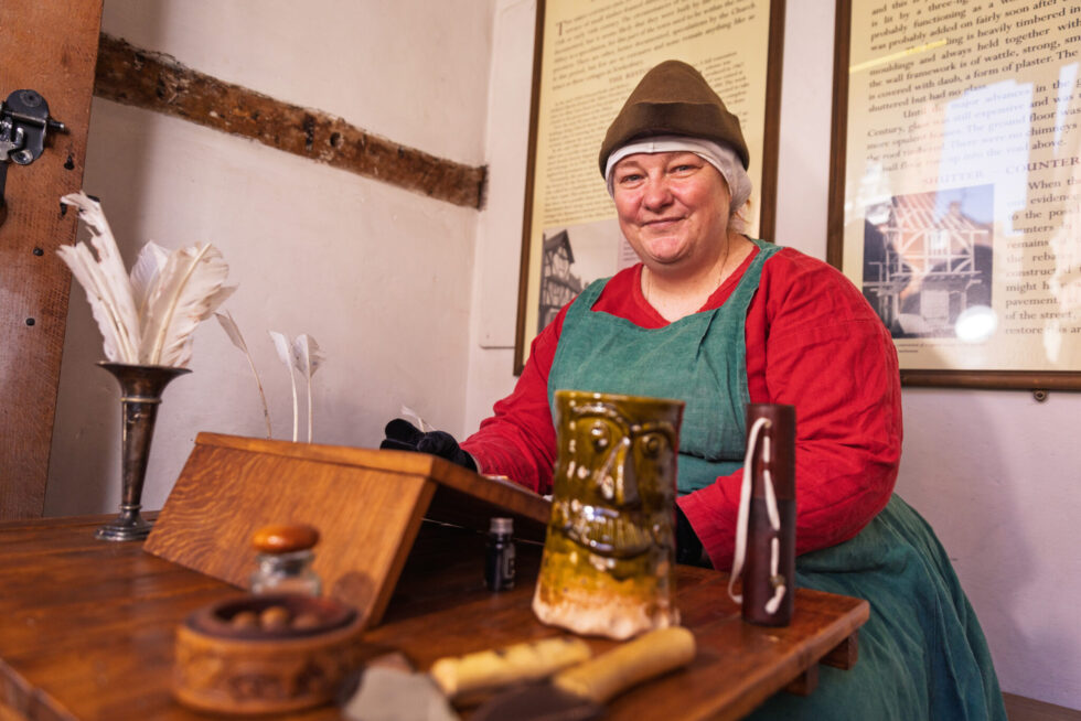A woman dressed in medieval dress sitting at a table with a quill and jar - House of Hound and Falcon