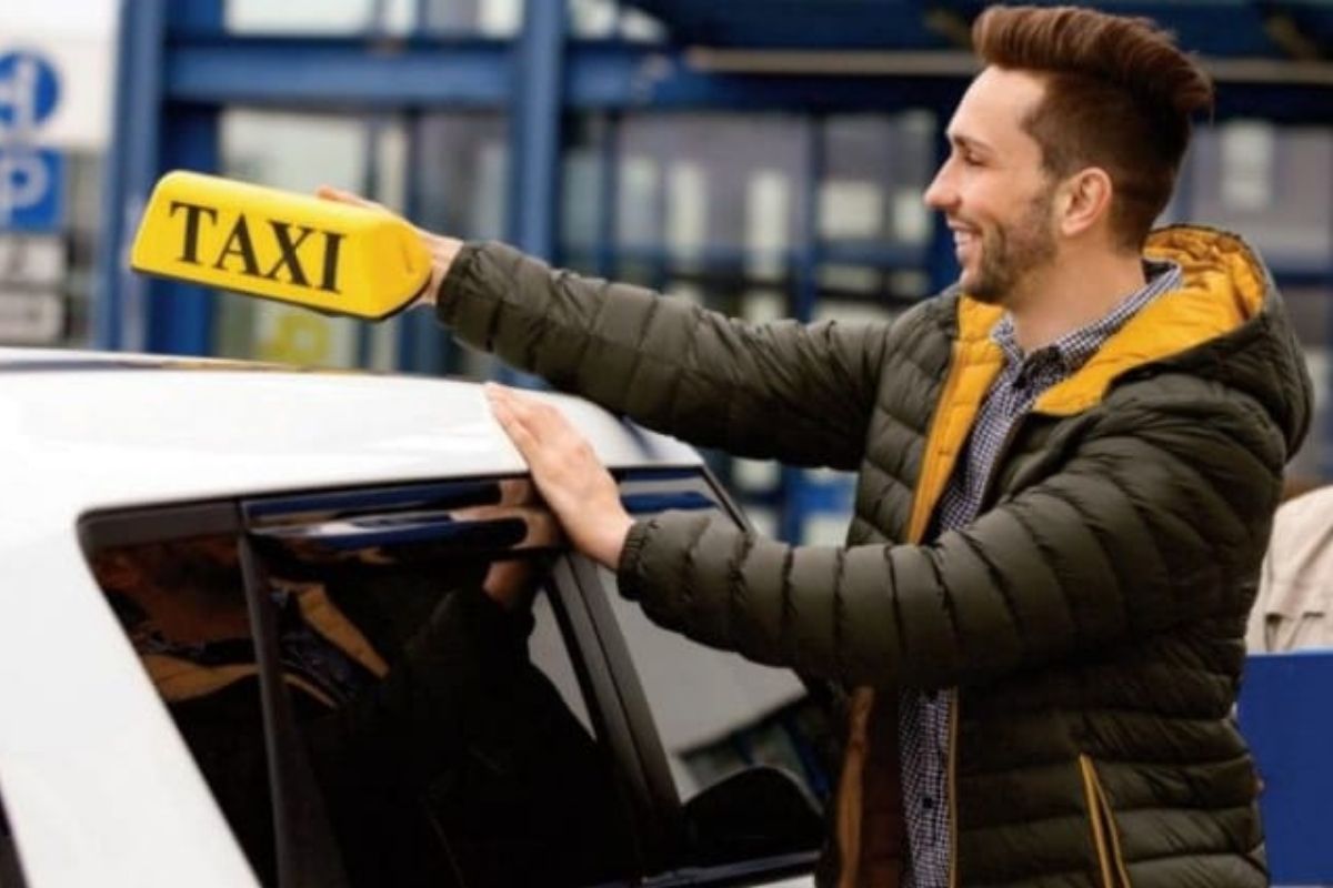 Photo of a man in a coat with a yellow hood, putting a yellow taxi sign on top of a white car.
