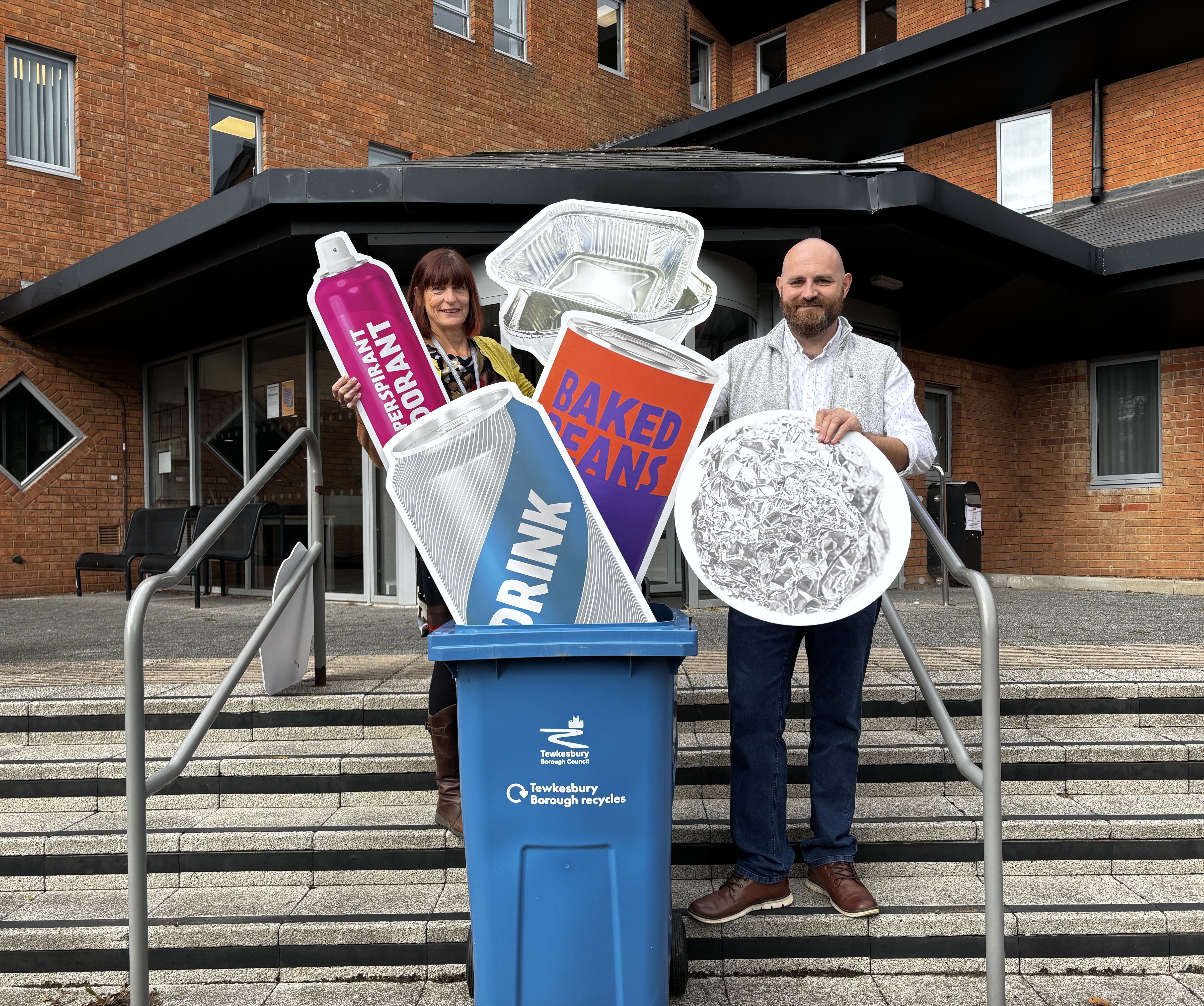Cllr Murray Stewart - Lead Member for Environmental Services and Kate Cole - Waste Services Officer, stand next to a blue recycling bin with large cut outs of metal recyclable objects